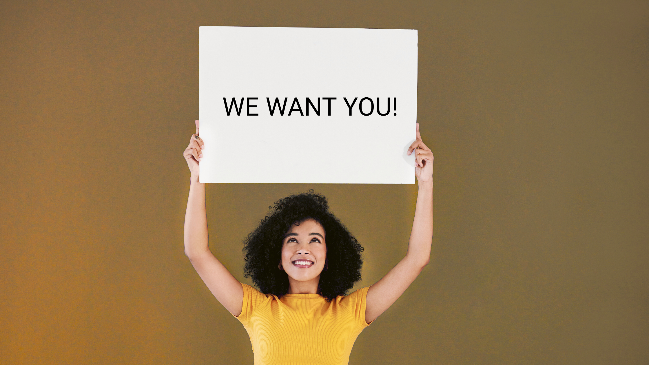 We Want You! Smiling woman in yellow shirt holding a white sign that says "WE WANT YOU" against a plain background.