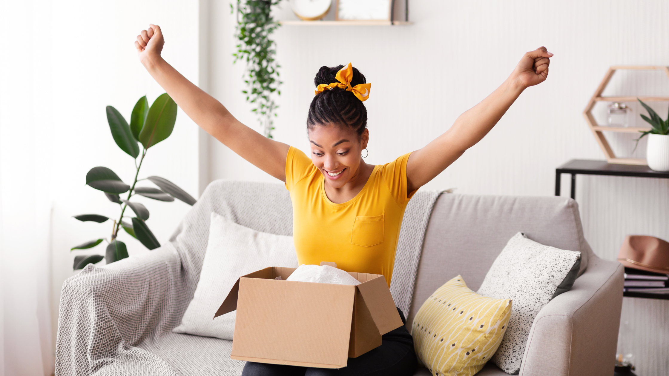 Your First PR Package Moment Excited woman in a yellow shirt raising her arms in celebration while opening a package on her couch.