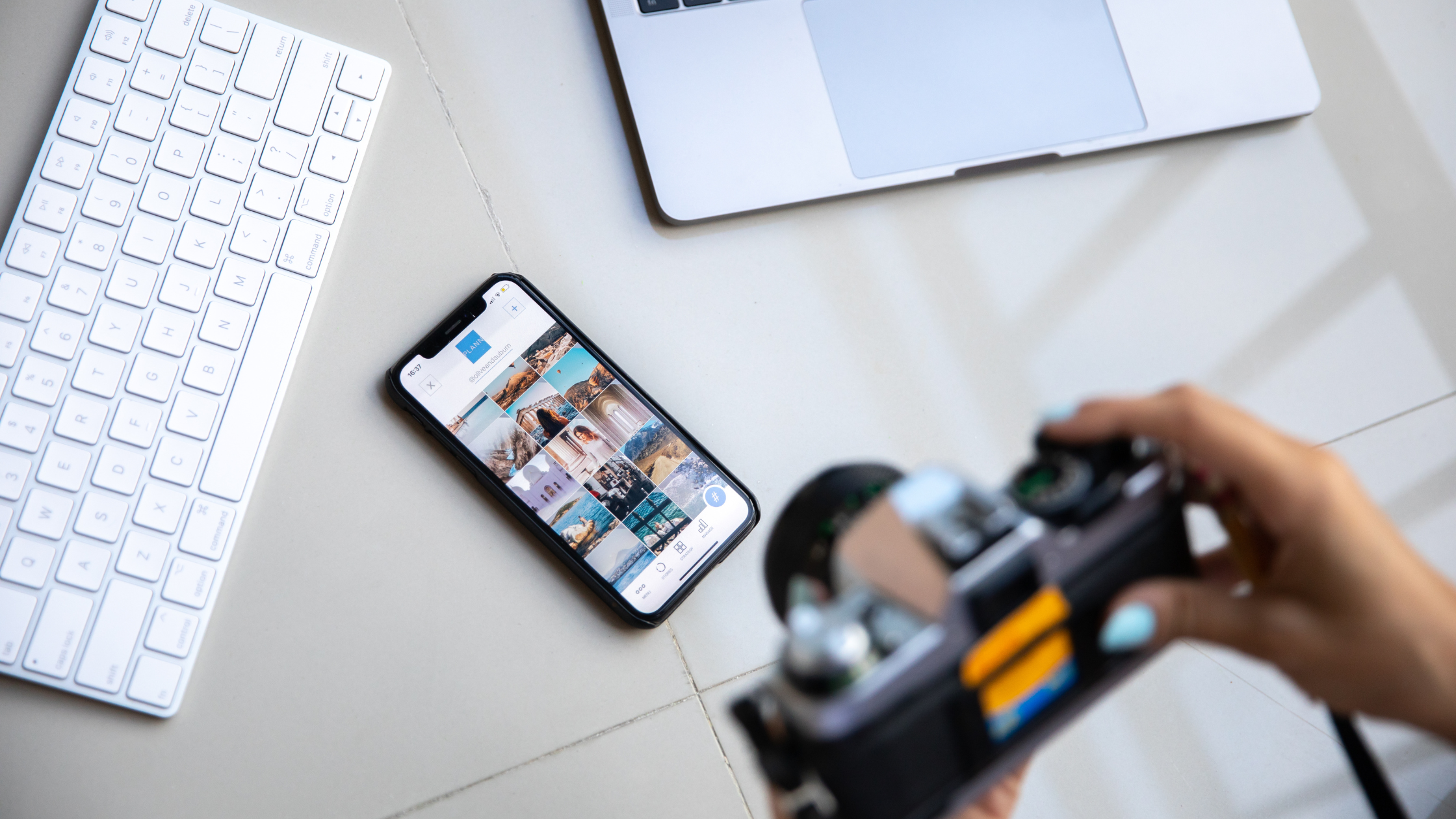 Content Planner Reviewing Instagram Feed Before Upload Person holding camera with a smartphone open to an Instagram grid on a desk with laptop and keyboard