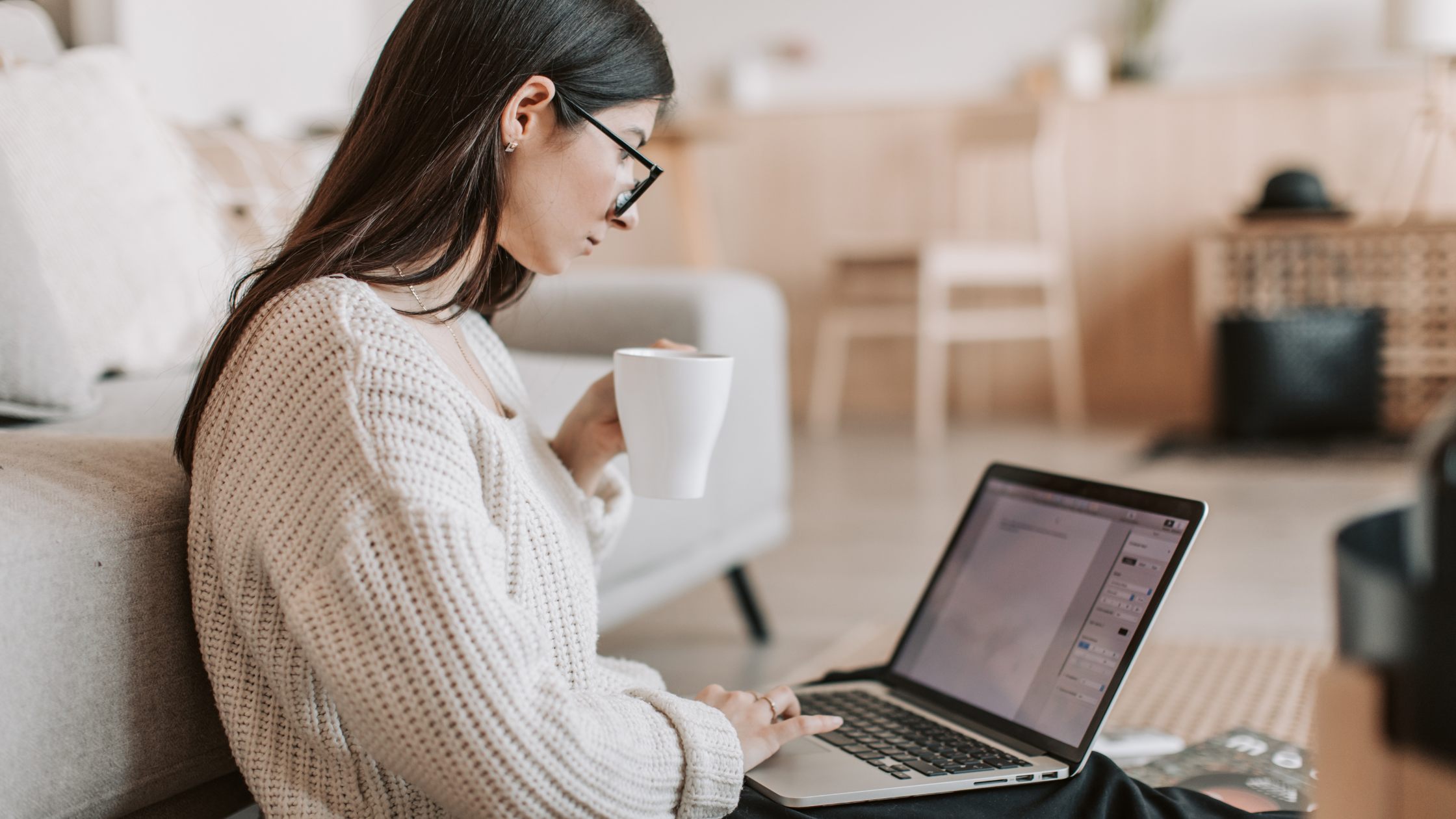 Woman working on laptop with coffee mug at home Woman in glasses working on a laptop at home while holding a coffee mug