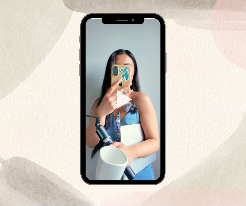 Woman in a blue dress taking a mirror selfie holding a phone, microphone, laptop, and coffee mug.