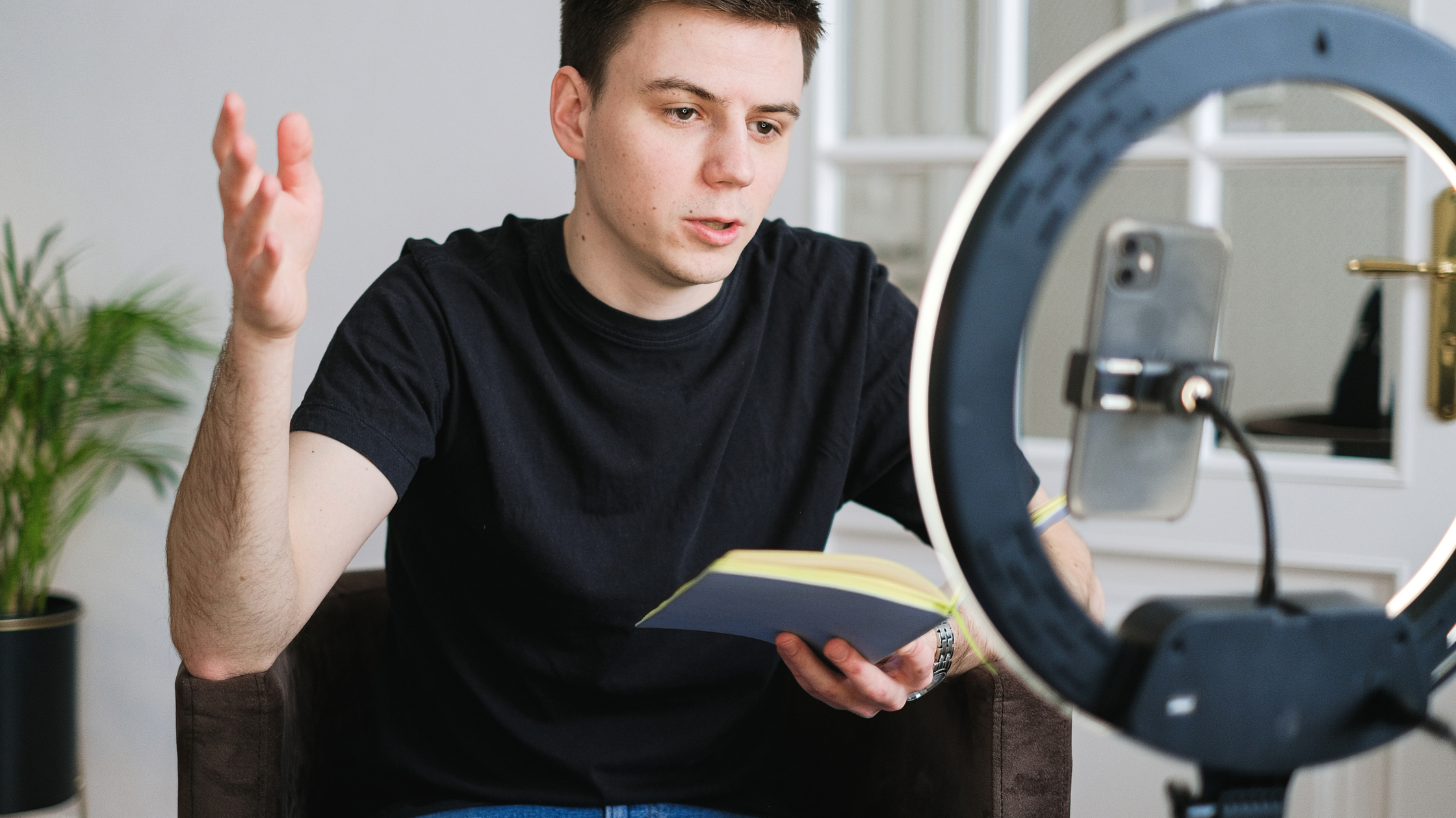 Young male creator recording video content with a smartphone and ring light while holding a notebook.