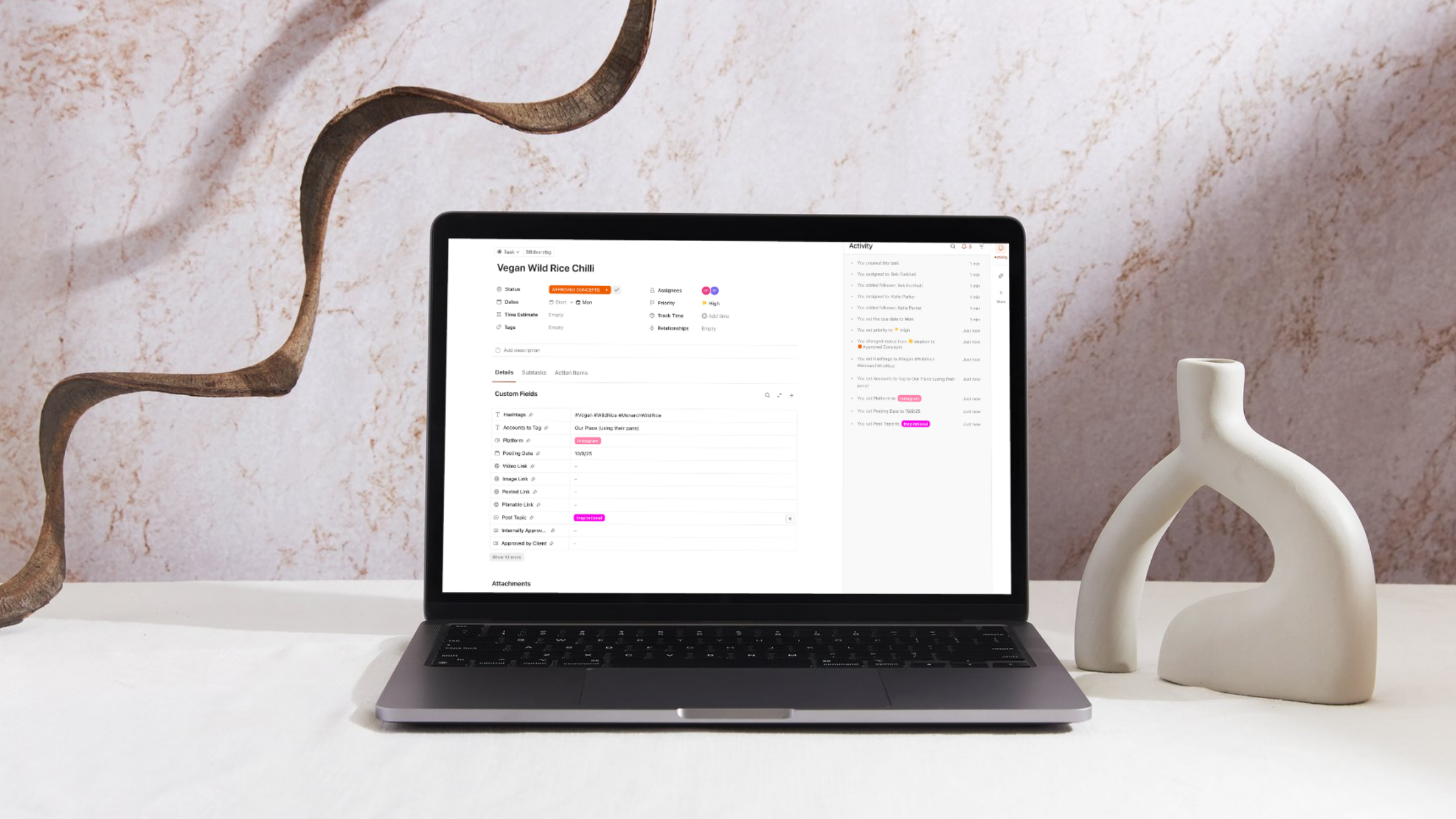 Laptop displaying a ClickUp project management dashboard with a content workflow task for social media scheduling, placed on a minimalist desk with sculptural décor.