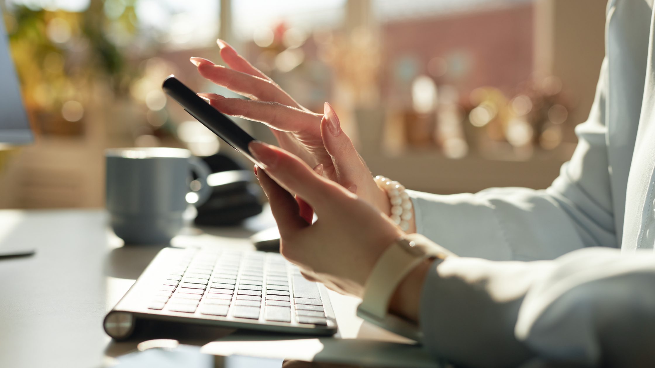 Close-up of hands holding a smartphone above a keyboard and coffee mug, symbolizing mobile communication and real-time social media management.