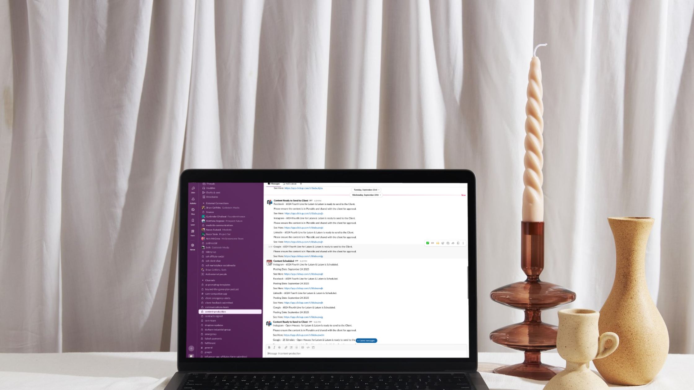 Laptop screen showing Slack workspace with client content approval messages, placed on a styled desk with candles and ceramic vases.