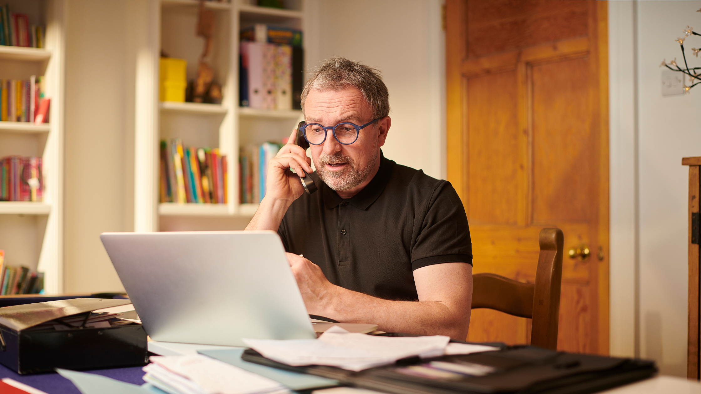 Man working from home, talking on the phone while reviewing business documents on his laptop.