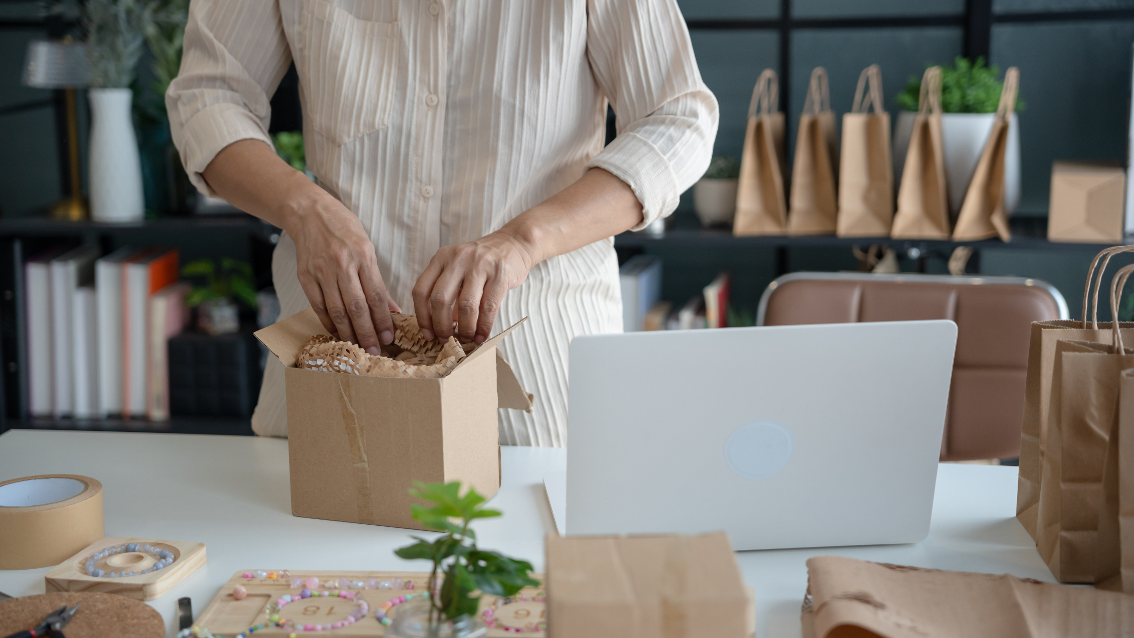 Small business owner packing products into a shipping box at a desk with a laptop.