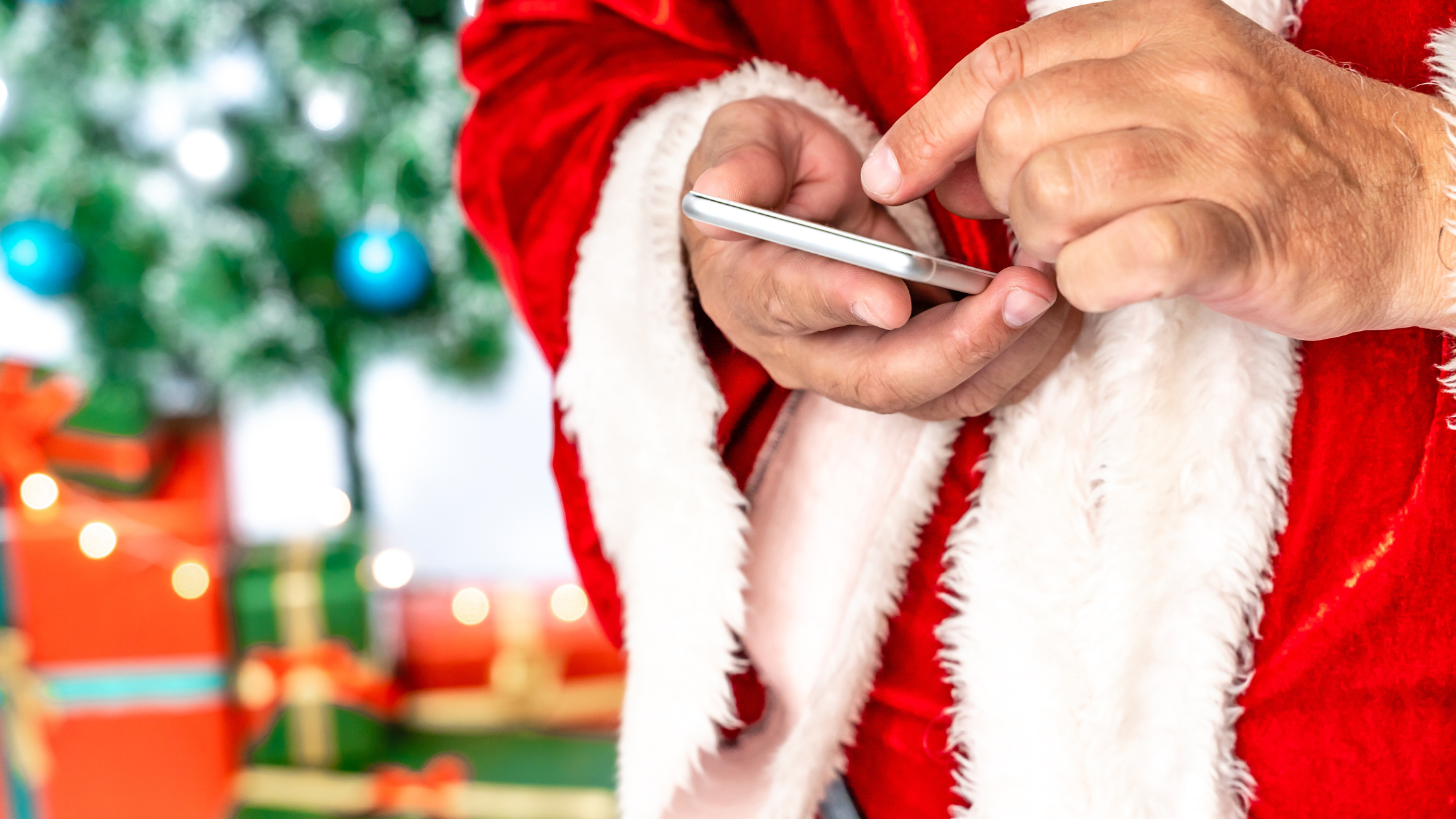 Person wearing a red Santa-style jacket using a smartphone with Christmas gifts and a decorated tree in the background.