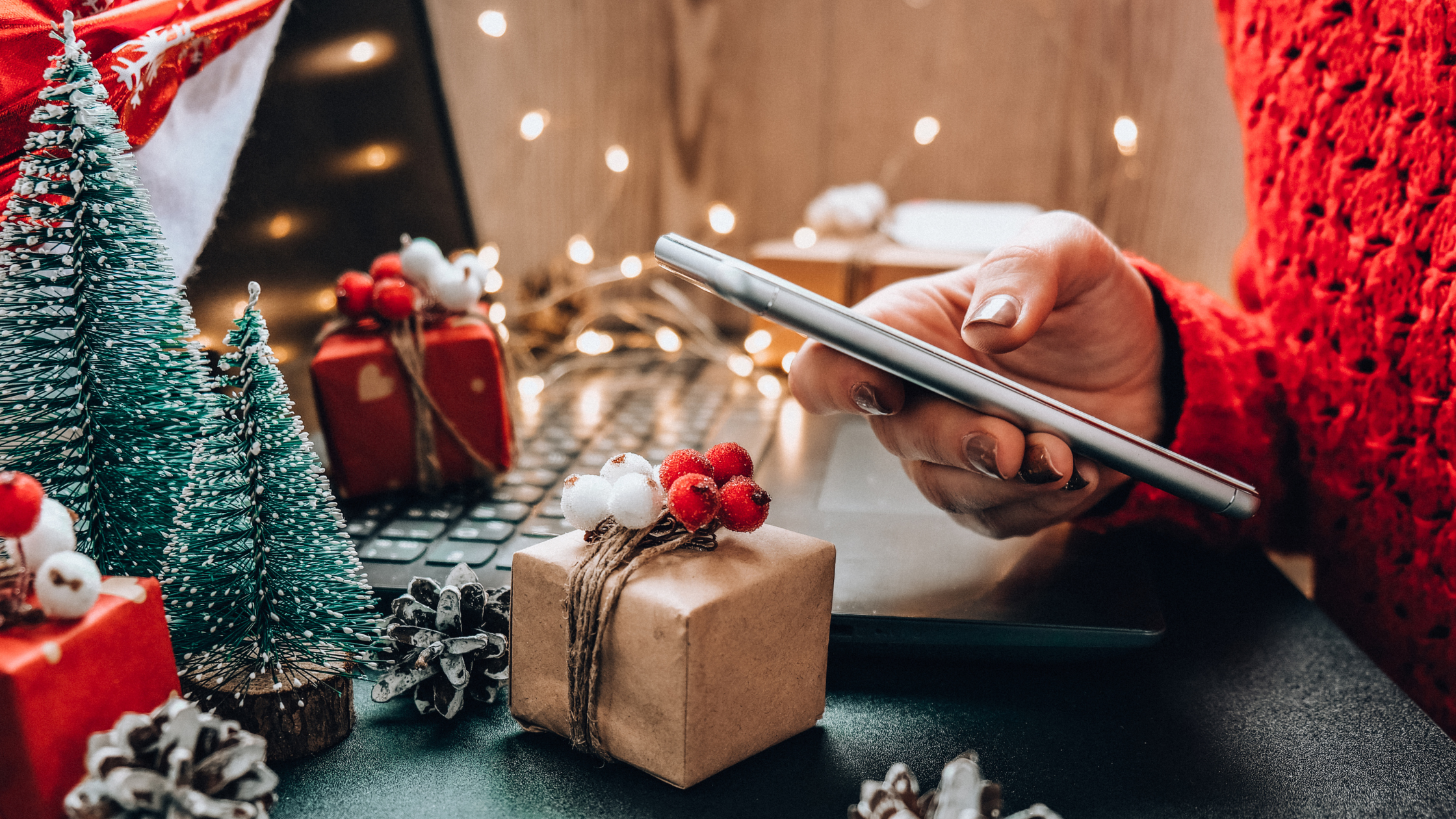 Hand holding a smartphone at a holiday-decorated desk with mini Christmas trees, wrapped gifts, and warm lights.