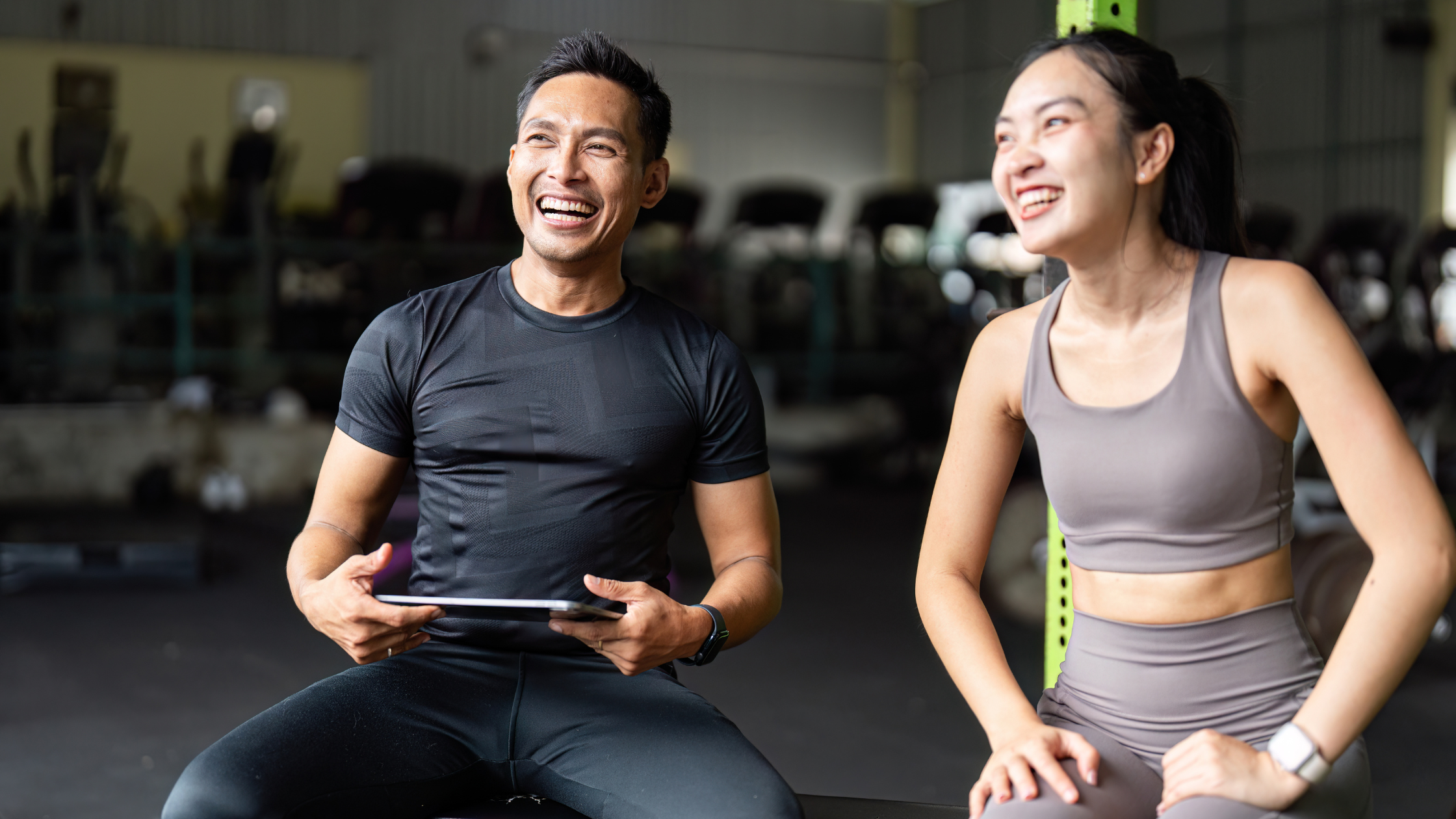Two people sitting in a gym laughing after a workout, showing positive energy and connection.
