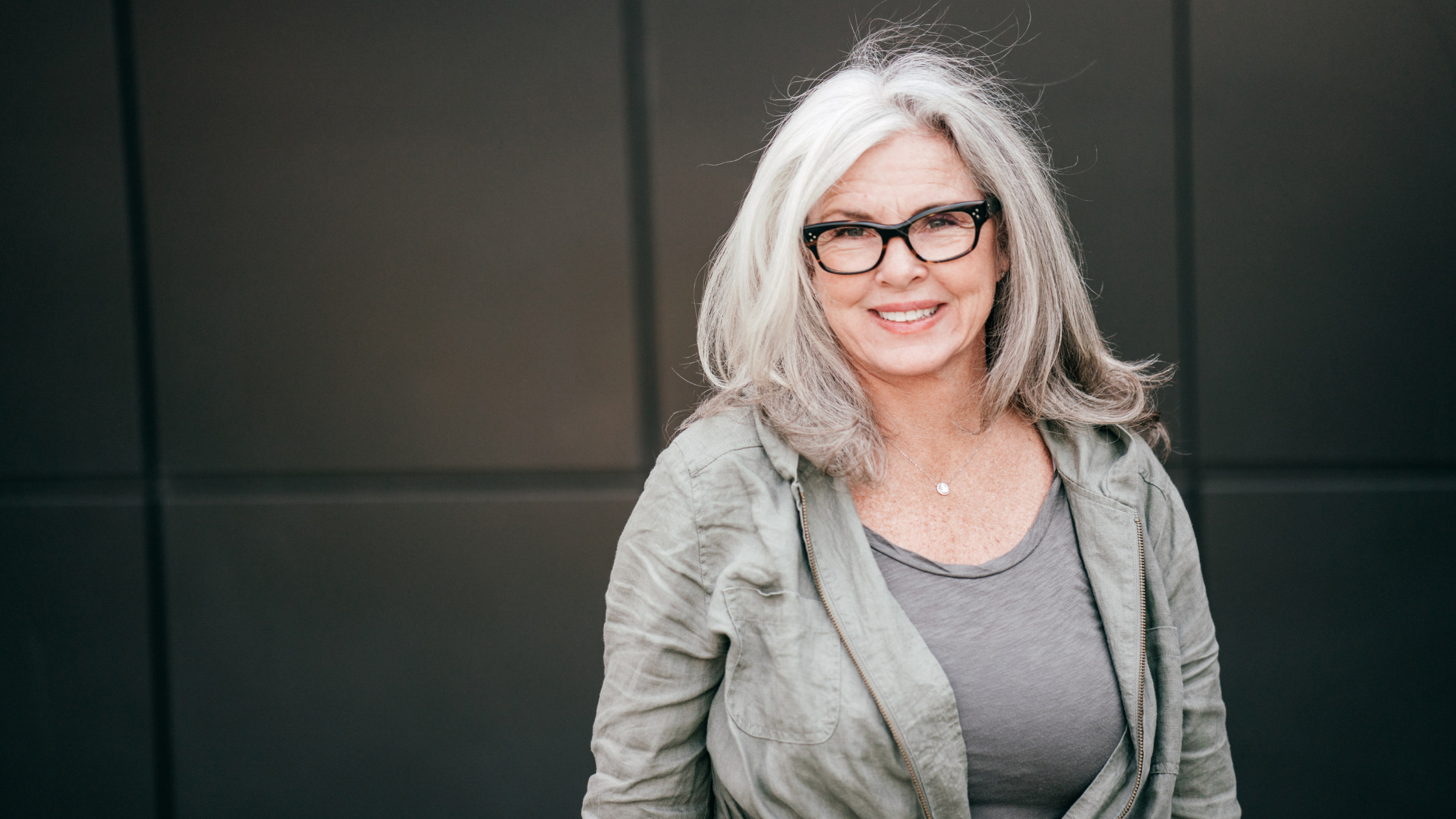 Confident woman over 60 with grey hair and glasses standing against a modern dark background