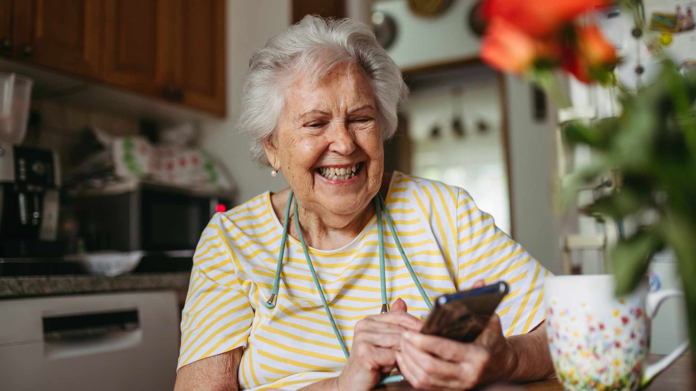 Smiling elderly woman using smartphone in cozy kitchen, representing social media engagement across generations