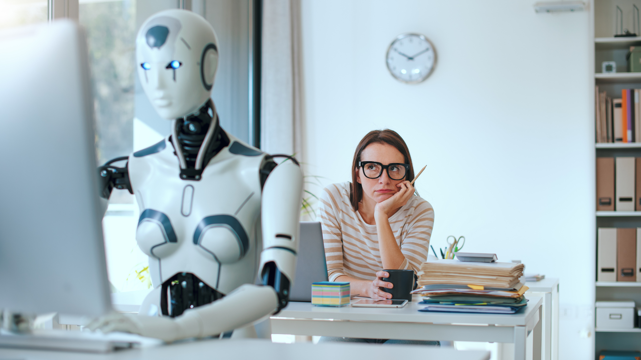 Humanoid robot working on a computer while a frustrated office worker sits behind a desk with paperwork