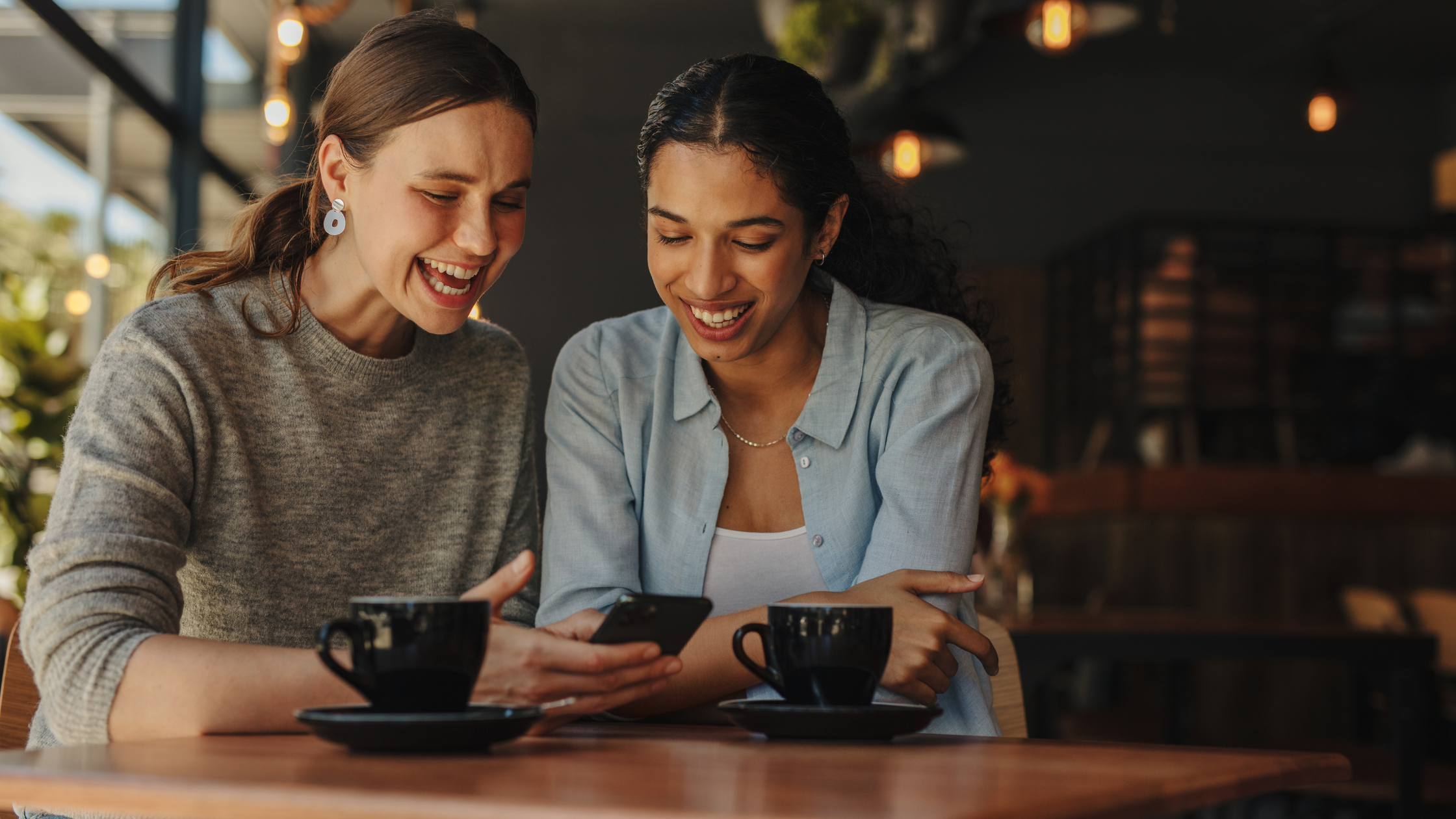 Two women laughing together at a cafe while looking at a smartphone, representing social sharing and authentic creator content that drives brand awareness through micro influencer marketing and product seeding.