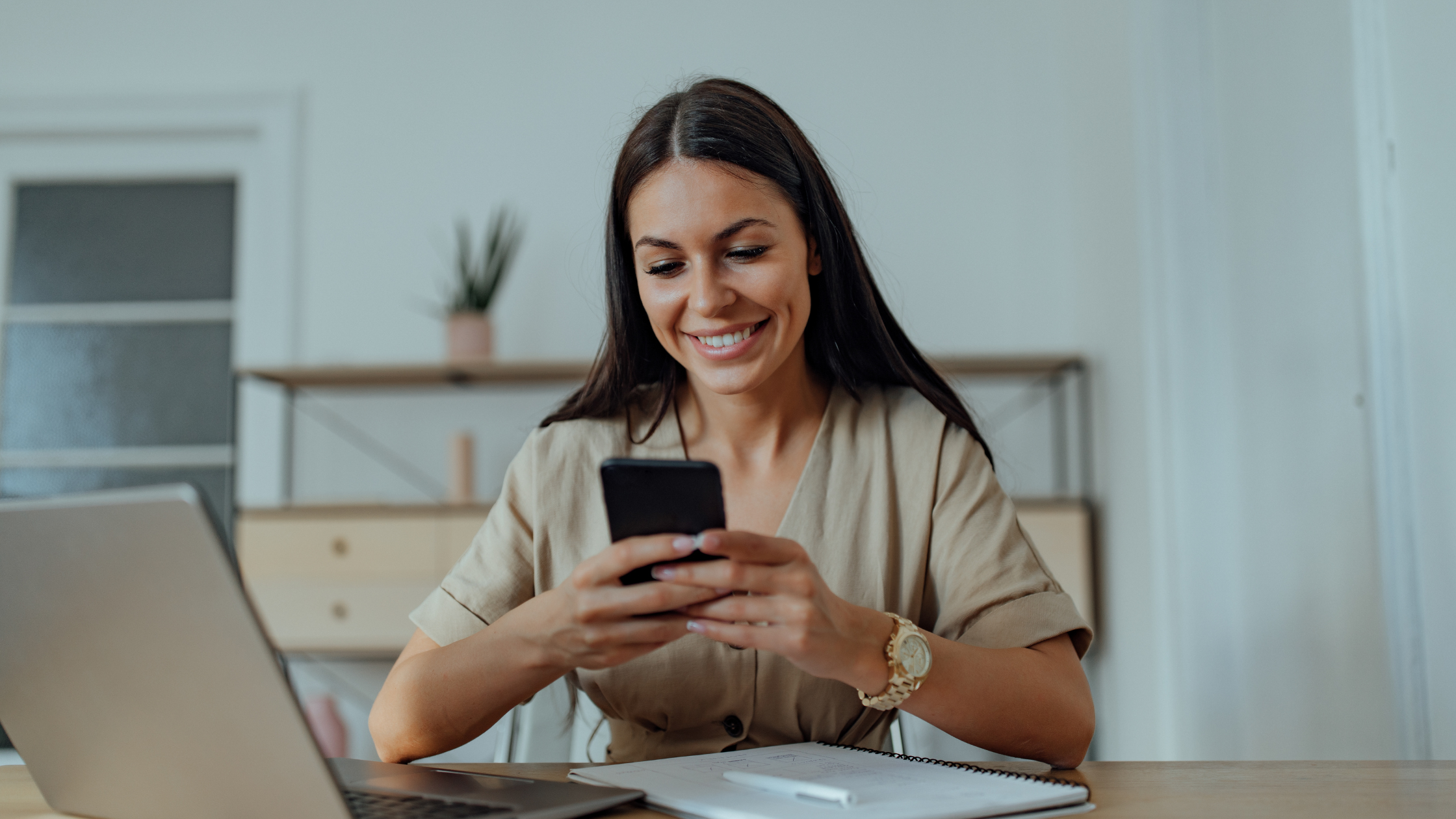 Smiling woman looking at her smartphone at a desk with a laptop and notebook, representing a brand marketer or creator engaging with micro influencer marketing content and product seeding campaign results.
