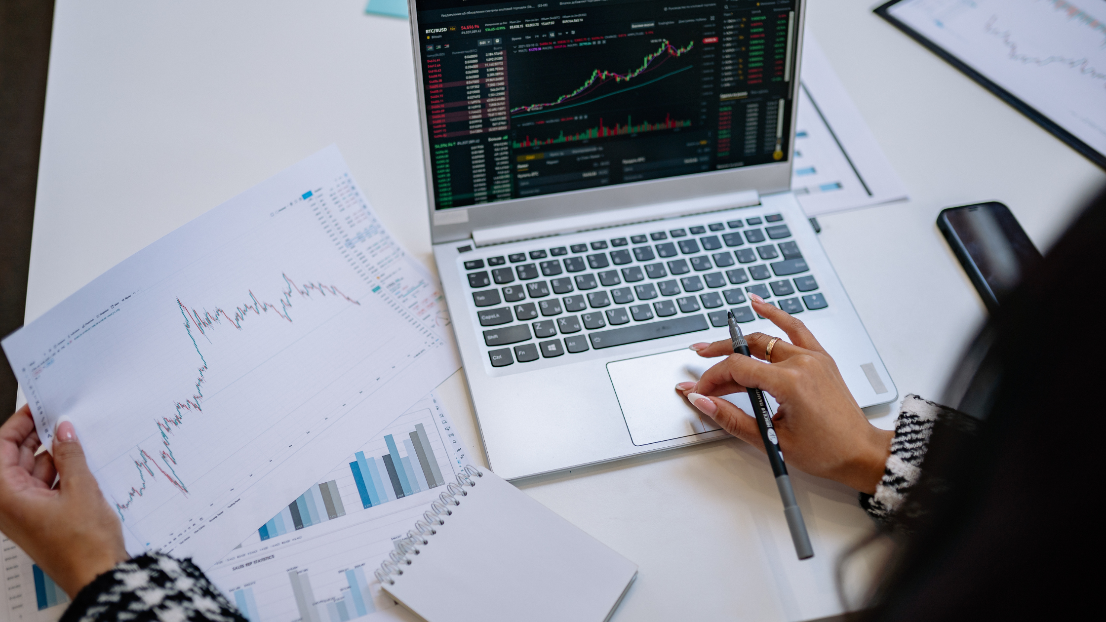 Person reviewing trading charts and performance data on a laptop and printed reports