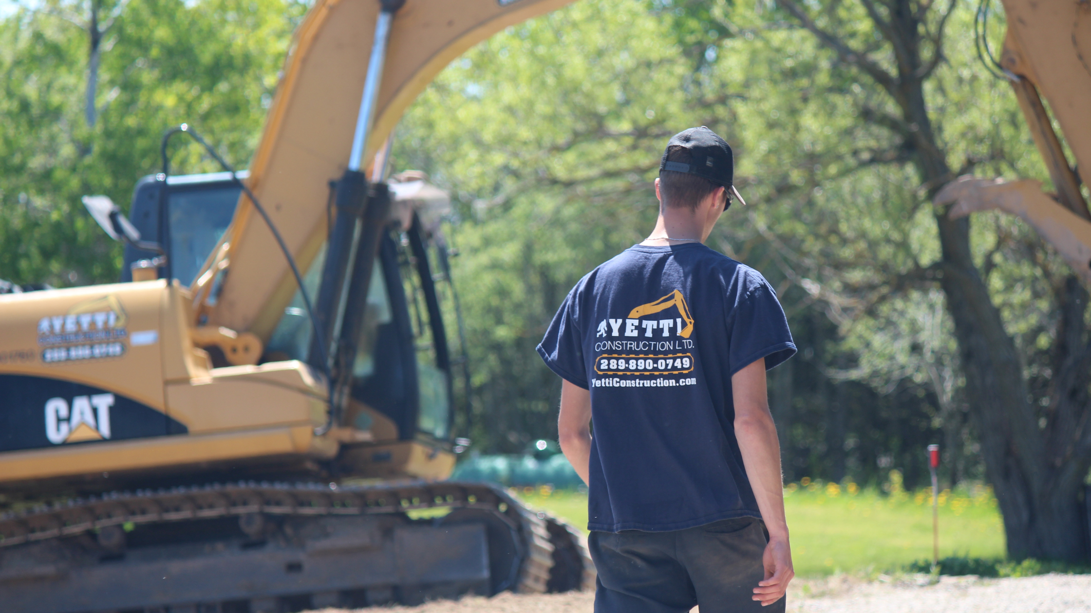 Yetti Construction employee in a branded t-shirt standing near a CAT excavator on a job site