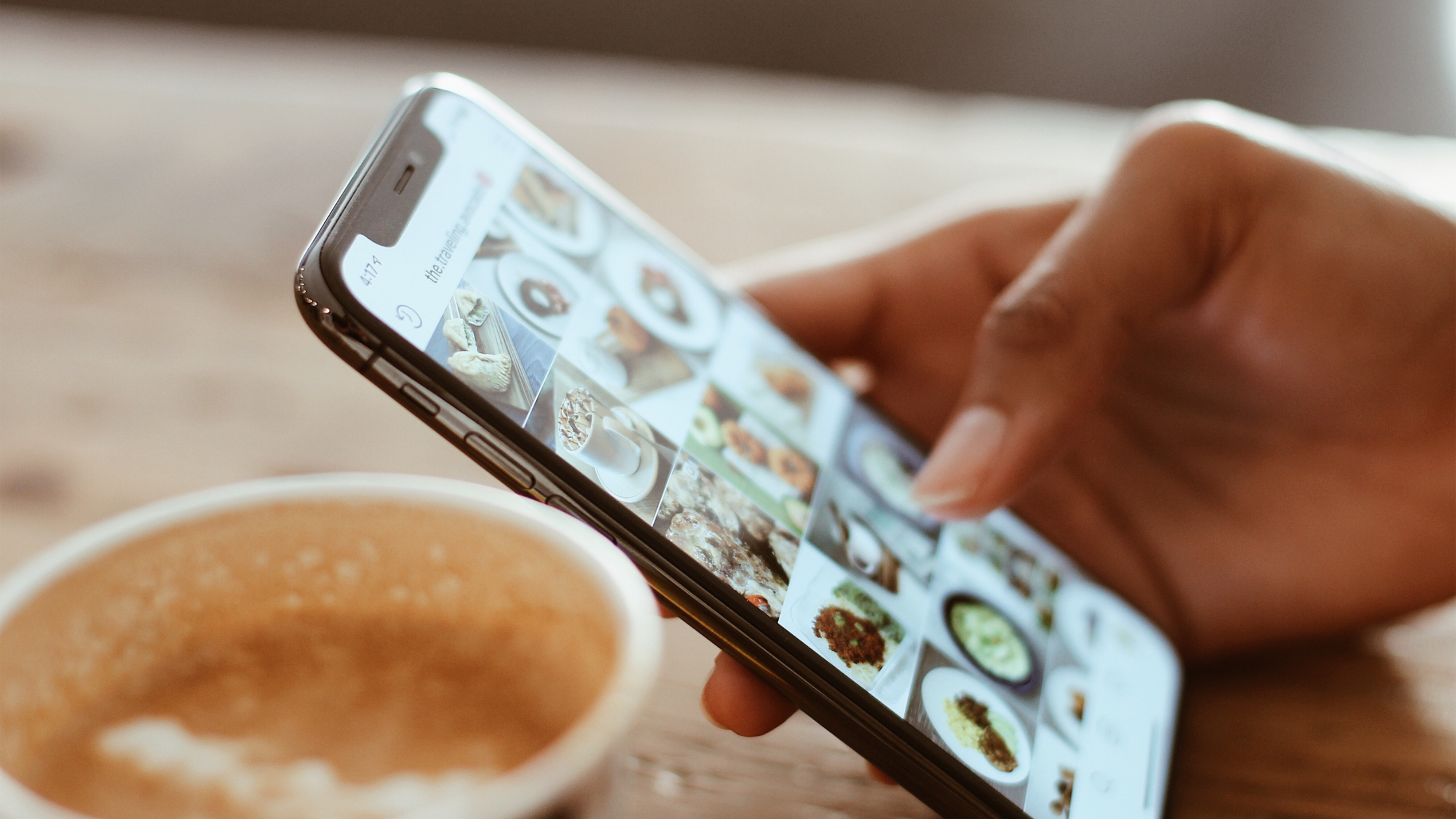 Hand holding a phone scrolling a crowded grid of Instagram posts next to a cup of coffee