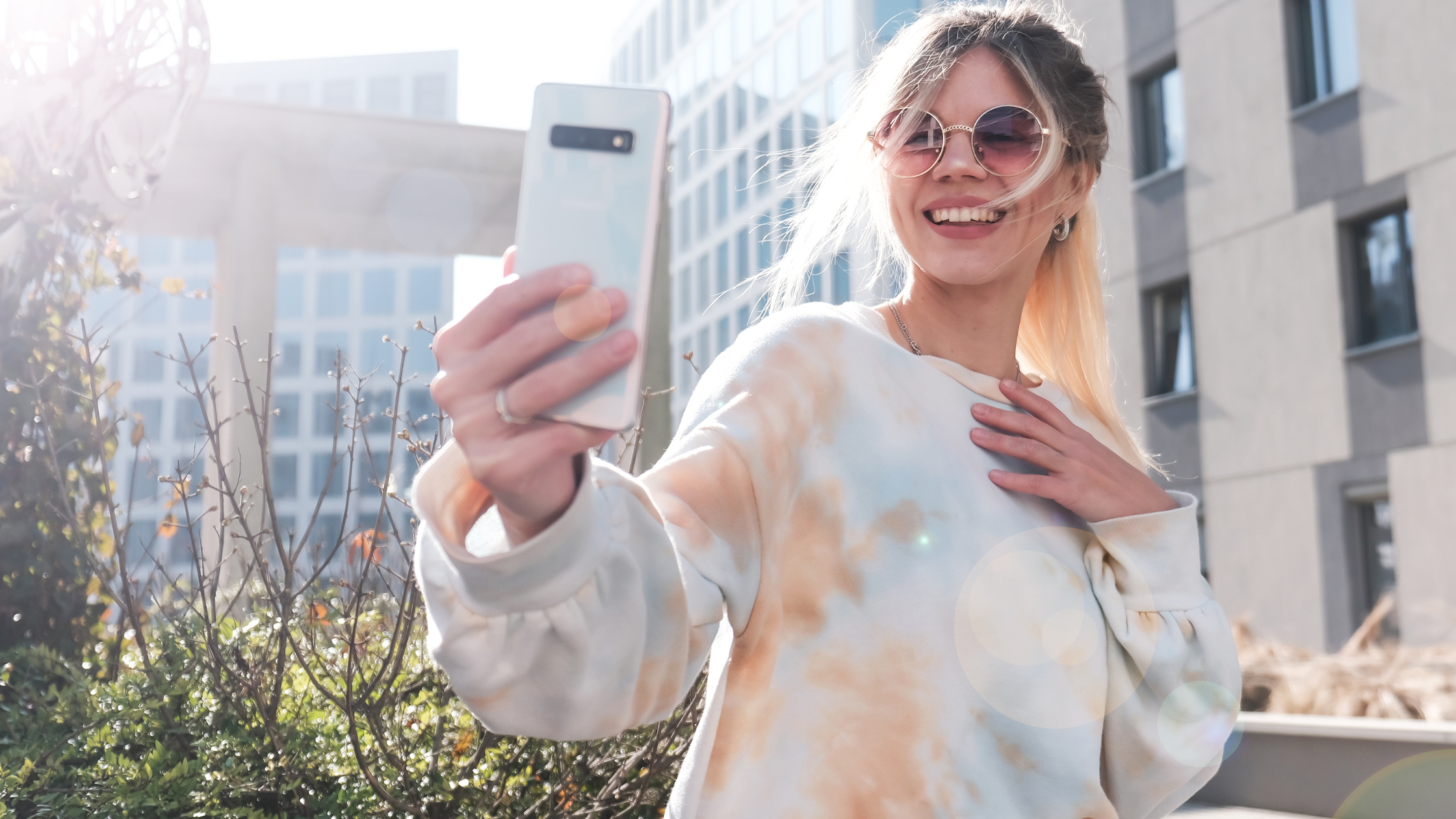 Smiling woman in round sunglasses and a tie-dye sweatshirt taking a selfie outdoors in the city, representing real, relatable creator content