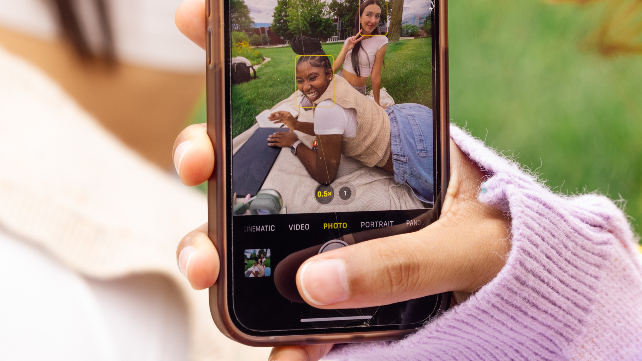 Hand holding a phone taking a photo of two smiling young women on a grassy lawn, representing relatable user-generated content and product seeding