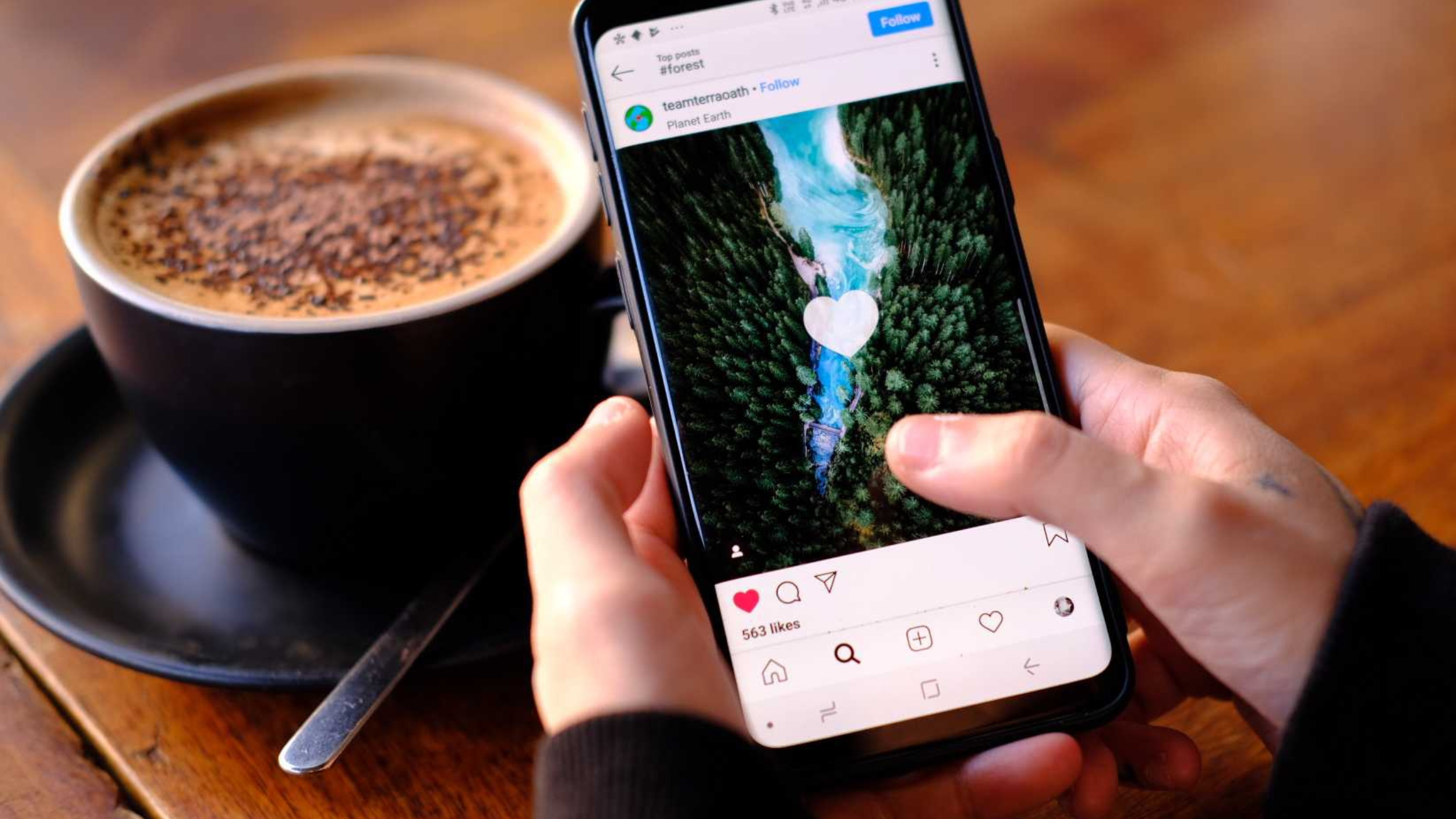 Hands holding a phone double-tapping to like an Instagram post next to a cup of coffee on a wooden table