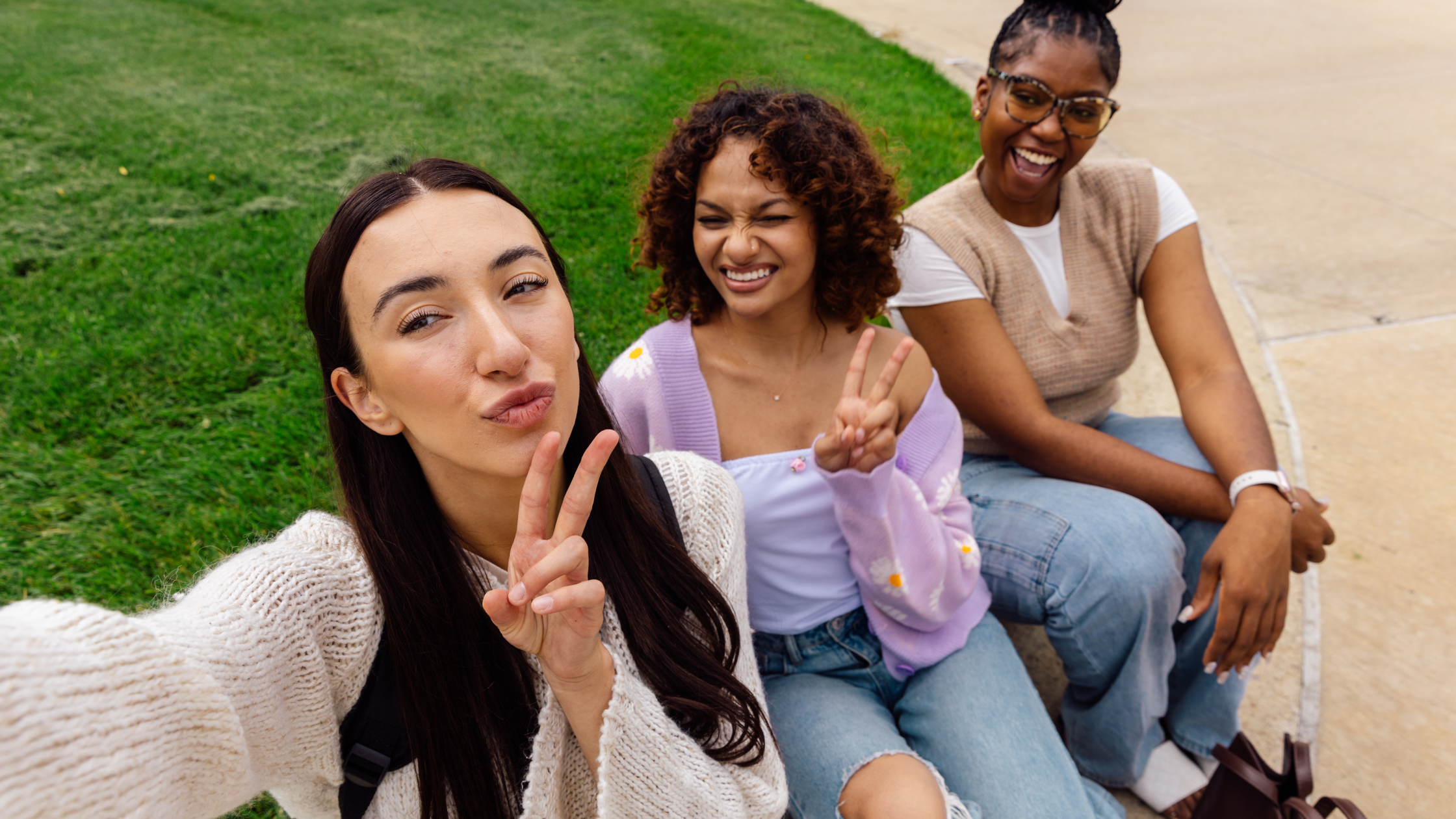 Three smiling young women taking a selfie together on a grassy campus lawn, representing creator content for student housing ads