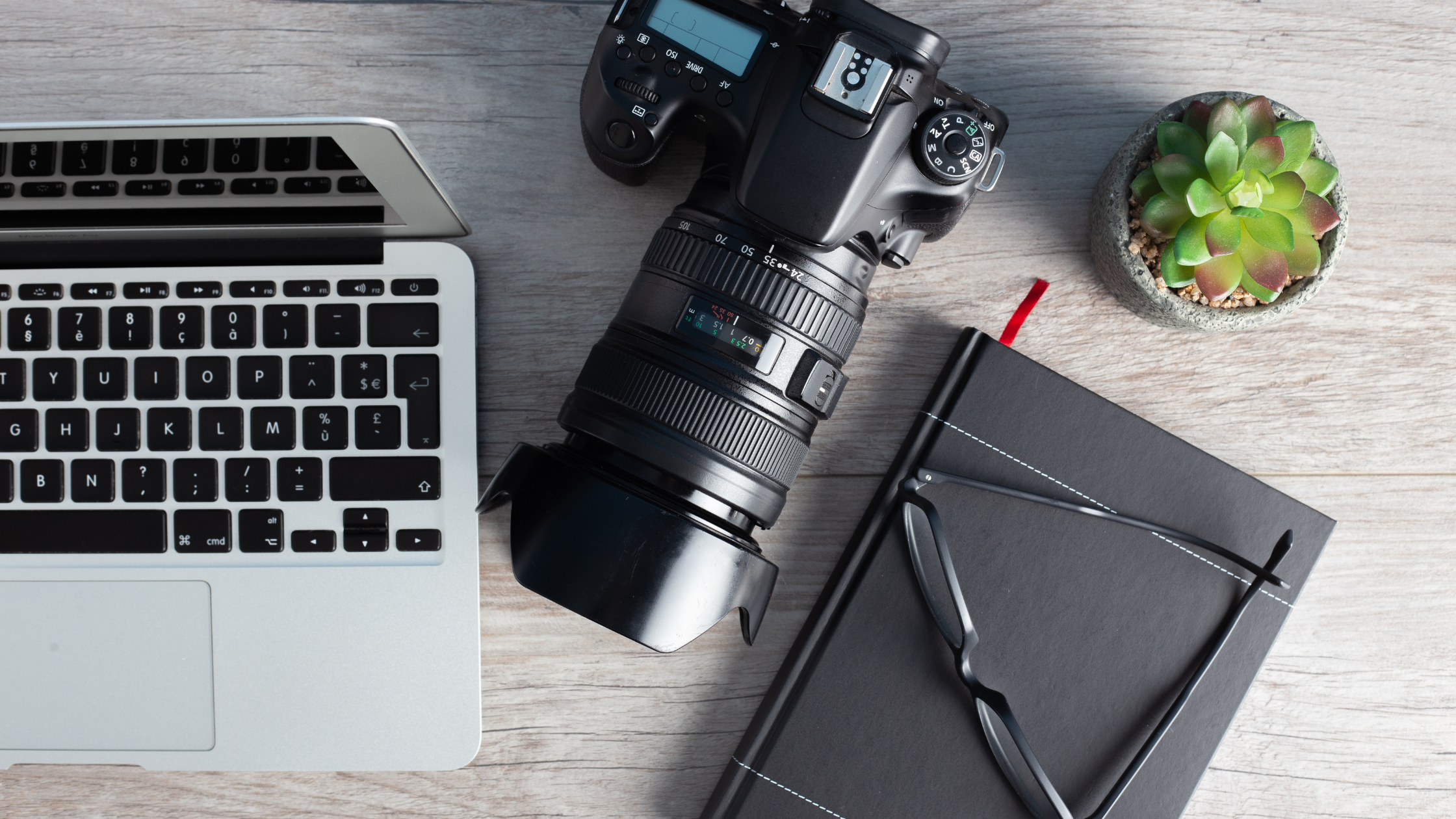 Overhead view of a laptop, DSLR camera, black notebook and glasses on a wooden desk, representing creator content planning